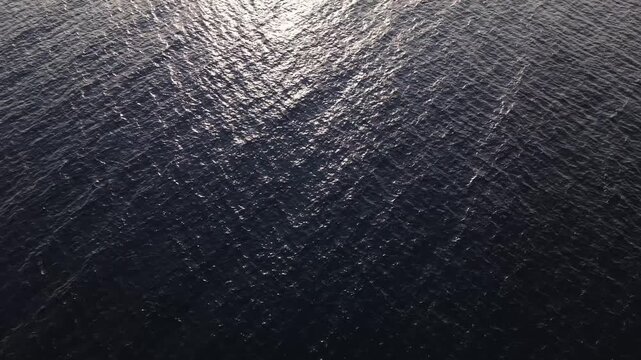 Overhead drone footage of ocean water with bright sunlight glinting across gentle waves, creating a dynamic pattern of sparkling reflections on the surface.