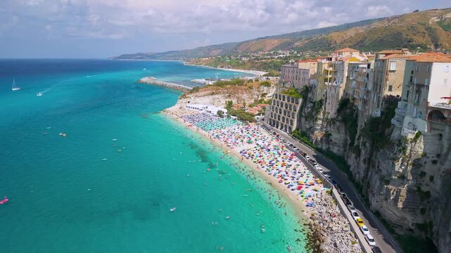 Tropea Calabria Italy 4k drone view of historic clifftop houses and sandy beach. Flyby along vertical rock wall turning to frontal view of pastel homes, colorful umbrellas establishing shot