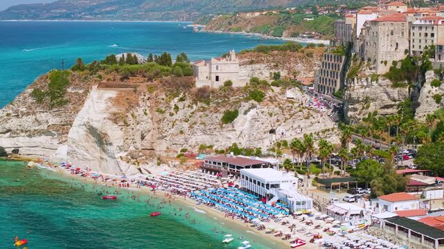 Tropea Calabria Italy 4k drone 70mm view of Santa Maria dell Isola sanctuary. Flythrough gap between rock and clifftop town reveal beach umbrellas, telephoto compression broll establishing shot
