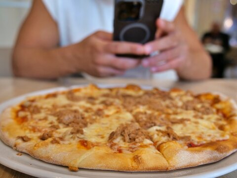 Close-Up View of Freshly Baked Crusty Tuna Pizza with Blurred Person Taking Photo using Smartphone in Background