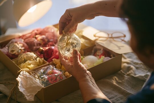Vintage christmas ornaments being lifted from a cardboard storage box by an elderly hand, a tender scene for heirloom and nostalgia