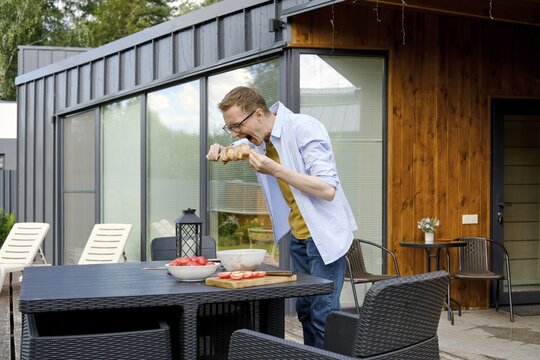 A middle-aged man stands at a patio table of tiny house and playfully bites marinated pork meat on skewer