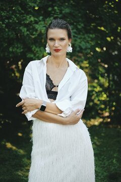 A female model stands outdoors in a lush green setting, wearing a white fringed dress paired with a black lace top