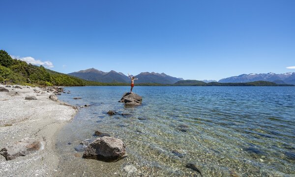 Young man stands on stone in water and stretches his arms in the air, Beach Frasers Beach, Lake Manapouri, Manapouri, South Island, New Zealand