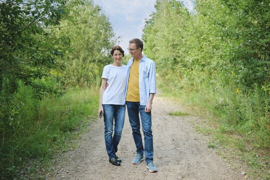 A middle-aged couple enjoys a stroll along a gravel path shaded by lush greenery. They smile at each other and have a leisurely conversation