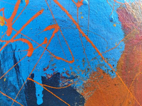 A bright blue painted background crossed by energetic orange paint splatters, drips and thin lines, with touches of red and yellow creating a lively abstract street-art composition.