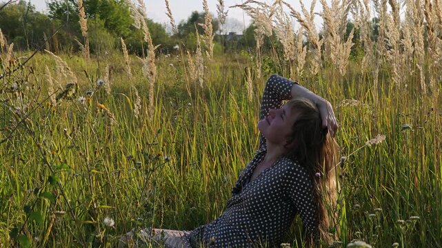 Woman sits in tall meadow grass and touches her hair at sunset.