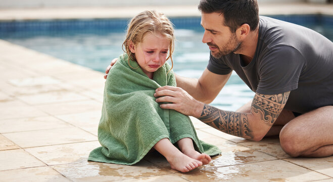 Man comforting a crying young girl wrapped in a green towel near a swimming pool