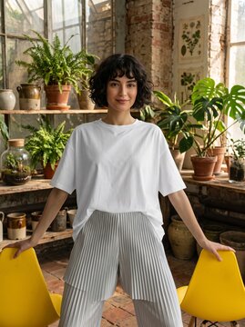 Woman wearing a white t-shirt standing inside a greenhouse filled with lush green potted plants and rustic wooden shelves.