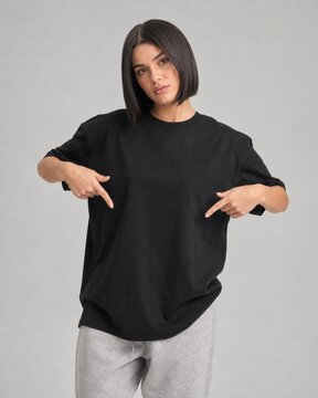 Brunette woman with short dark hair wearing a black t-shirt and gray sweatpants pointing down at her torso.