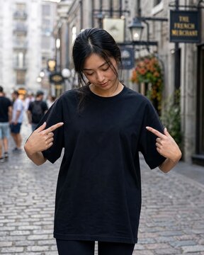 Woman wearing a plain black t-shirt standing on a cobblestone European street pointing down at her chest.