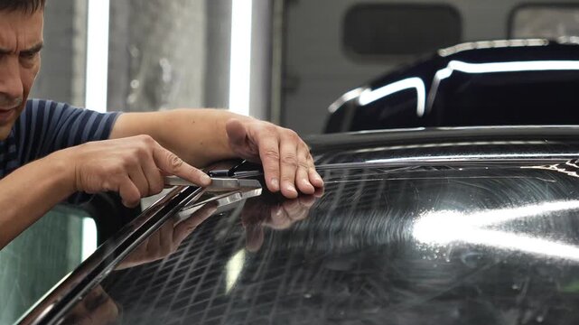 Professional auto detailer cutting film with a sharp knife on a car rear window glass in a vehicle service station workshop, window tint installation process. Male worker install dark window tinting. 