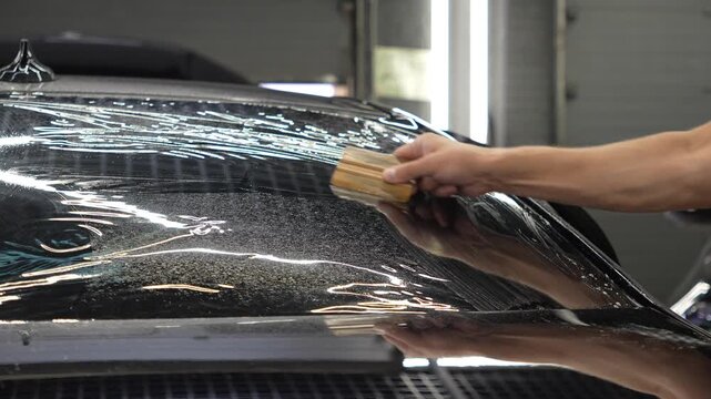 Professional auto detailer smoothing film with a squeegee tool on a car rear window glass in a vehicle service station workshop, window tint installation process. Male worker install dark tinting. 