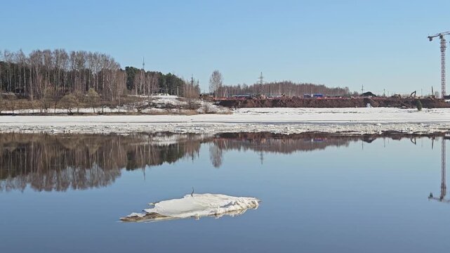 Floating ice drifts on calm, reflective river surface. Snowy banks frame distant trees and construction cranes. Clear blue sky mirrors the scene below. Still water reflects sky, trees