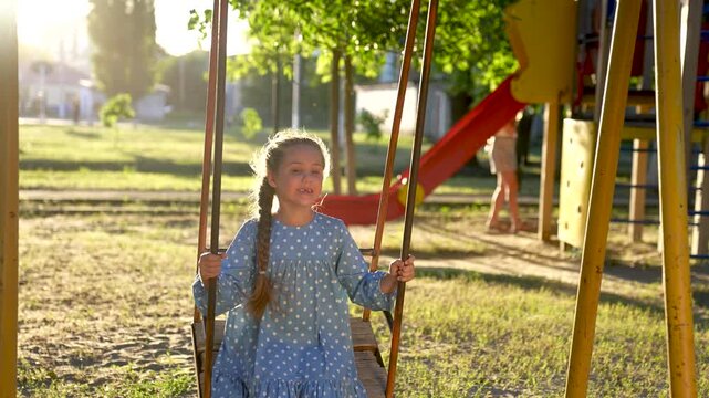 school girl swings on swing. concept of a happy childhood and loving family lifestyle. little child in blue dress swings on a swing on the playground, sunset and park in the background