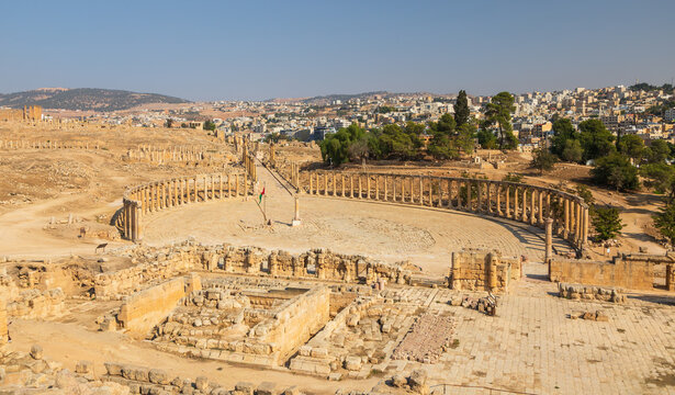 Aerial view of the ancient Roman Oval Plaza with its surrounding colonnades and historic stone ruins under a clear blue sky in Jerash, Jerash Governorate, Jordan.