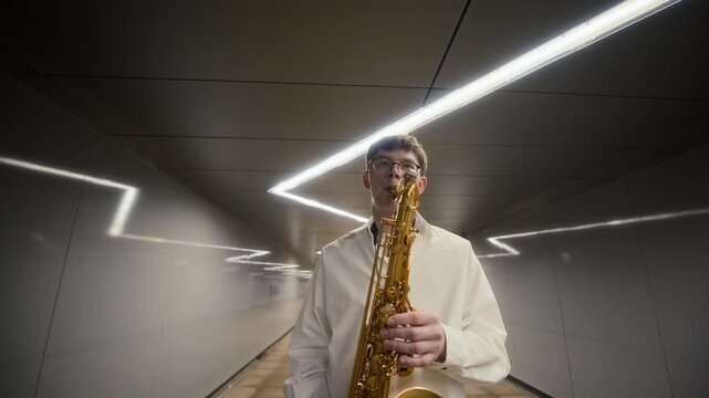 Caucasian young adult man playing saxophone in modern pedestrian tunnel from low angle. Visuals ideal for youth music education, urban culture campaigns, and creative talent promotions