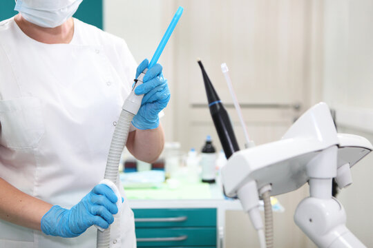 Dental worker in mask and blue gloves holds saliva ejector with hose. Clinic equipment background