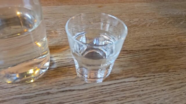 Clear glass bottle and shot glass sit on wooden table. Brown leather booth provides background texture. Contents show no visible liquid or labels. Setup suggests drinking or serving context