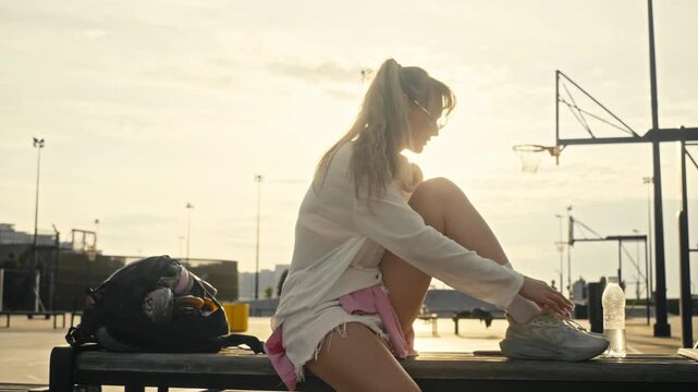Young woman ties sneaker laces after rollerblading in summer park. Tired skater rests on wooden bench packing sports gear into black backpack during beautiful warm golden hour sunset.