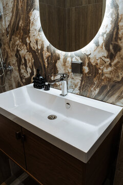 White rectangular bathroom sink with chrome faucet and mounted illuminated oval mirror on brown and beige marble patterned wall tiles.
