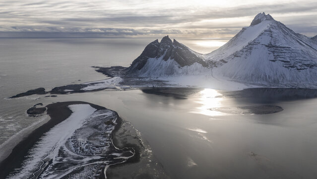 Aerial view of Vestrahorn mountain covered in snow during winter with reflective coastal waters and dramatic light in Hofn, Sveitarfelagid Hornafjordur, Iceland.