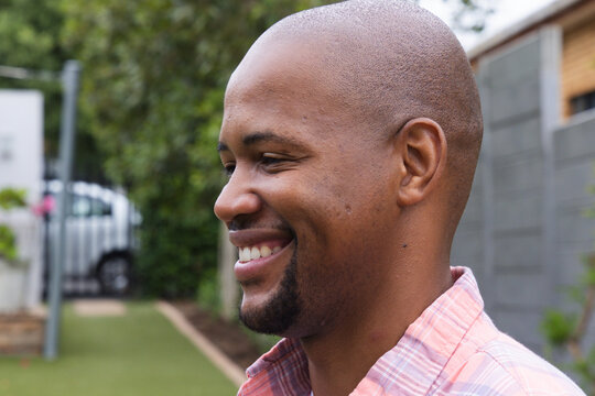 African adult man smiling and looking left in yard near path wearing collared pink plaid shirt