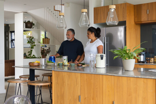 African American senior couple preparing fresh juice at kitchen island with blender and pitcher
