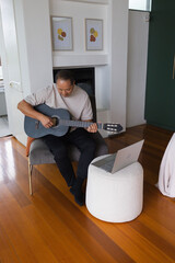 Fototapeta premium Senior Asian man playing black classical guitar in living room while referencing silver laptop