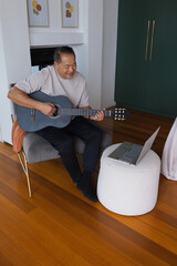Fototapeta premium Senior Asian man playing matte black classical guitar sitting on chair in living room with laptop