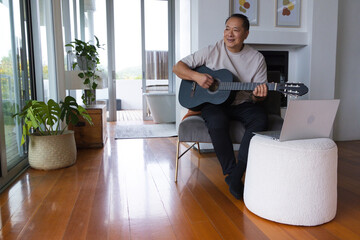 Fototapeta premium Senior Asian man playing guitar in living room wearing beige shirt, laptop nearby, copy space