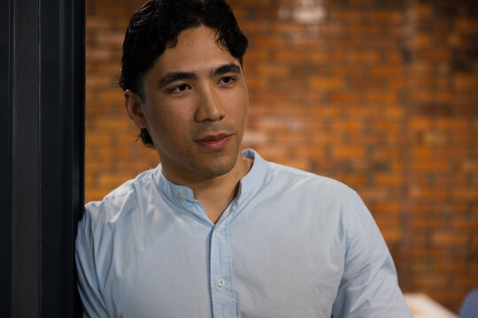 Asian man standing in brick-walled room near dark vertical frame at left, wearing light blue shirt