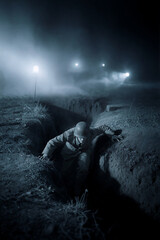 Fototapeta premium Soldier crawling out of a muddy trench at night. Vertical photo of a historical military battle with bright searchlights in thick fog. World War combat concept