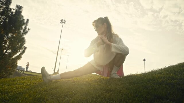 Low angle shot shows young woman stretching legs on grassy hill. Golden hour sunlight heavily backlights sportive female doing side lunge exercise near park skatepark ramp.