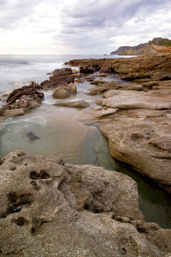 a rugged, weathered rocky shoreline with small tidal pools reflecting a partly cloudy sky and gentle waves rolling into the sea under a soft daylight. Argentiera, Sardinia, Italy