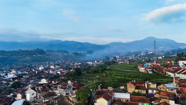 Aerial view of Ciwidey town featuring lush green rice terraces, dense residential rooftops, and misty mountains under a clear blue sky in Bandung, West Java, Indonesia.
