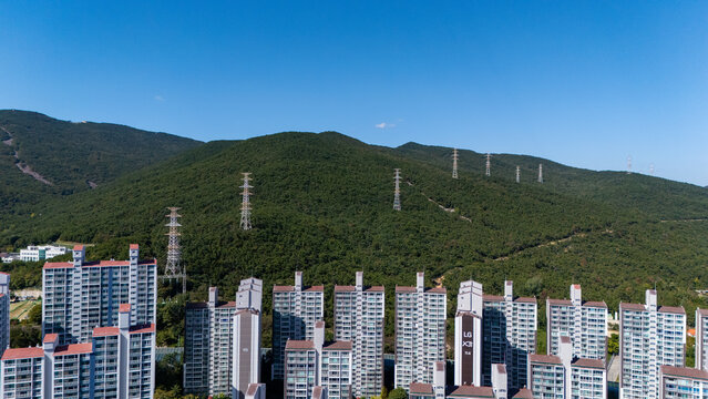 Aerial view of Jangsan mountain with high-voltage power lines rising above a dense residential apartment complex under a clear blue sky in Busan, Busan, South Korea.
