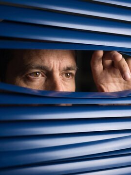 Man peeking through blue window blinds looking outside with curiosity
