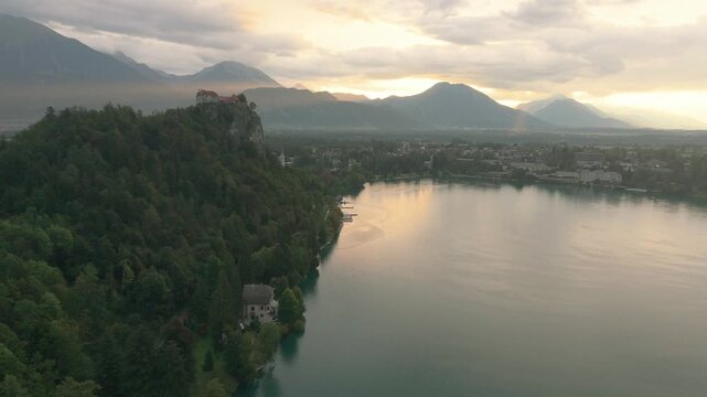 Aerial view of a medieval castle with red-tiled roofs and a stone tower perched on a dramatic rocky cliff, surrounded by lush forests, Bled lake and rolling hills in a picturesque European valley, Slo