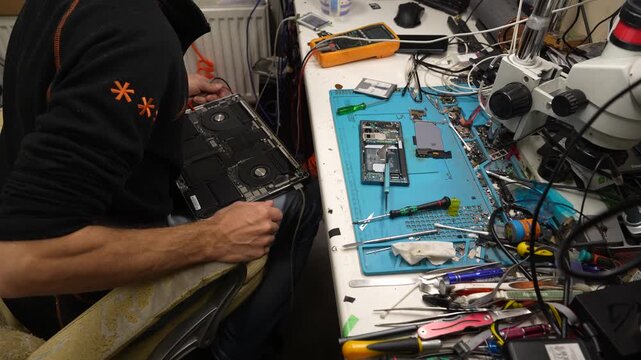Technician testing an opened electronic device with probes beside microscope and repair tools at a cluttered electronics workbench