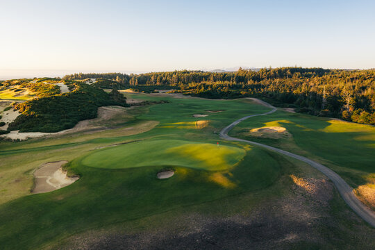 Aerial view of a golf course featuring rolling green fairways, sand bunkers, and surrounding coastal forest under soft golden hour light in Bandon, Oregon, United States.