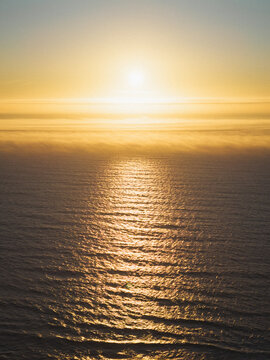 Aerial view of the Pacific Ocean at sunset with golden light reflecting on the water surface and a layer of low-hanging mist near the horizon in Bandon, Oregon, United States.