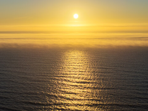 Aerial view of the sun setting over the Pacific Ocean with golden light reflecting on the water and a layer of mist on the horizon in Bandon, Oregon, United States.