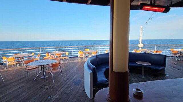 Ocean views from a ship deck cruise bar. Wooden floor, chairs, and tables. Blue sky, calm sea, distant horizon. Cabin view opens to sunlit deck. Bar stools and couches under shade