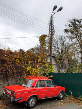 Kyiv, Ukraine - Nov 03, 2022: A vibrant red Lada sedan featuring a roof rack is parked on an unpaved street in the historic Kudriavets district, alongside a vine-covered wall and utility pole