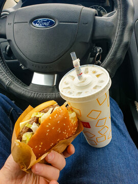 Kyiv, Ukraine - Mar 31, 2023: A person holds a fast-food burger, featuring a McDonald's disposable drink cup, inside a vehicle interior with a visible steering wheel
