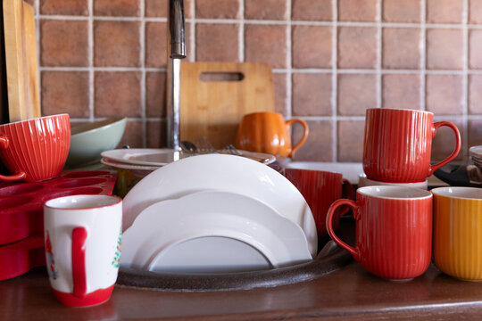 A busy home kitchen with colorful cups, plates and cookware arranged around the sink and countertop, capturing an ordinary moment of household life. The scene conveys domestic routine, unfinished chor