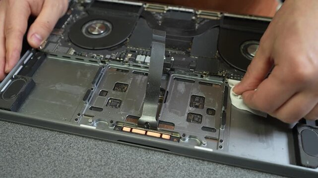 Technician cleaning inside an opened laptop with a cloth, revealing cooling fans, batteries, circuit boards, and internal components during electronic repair