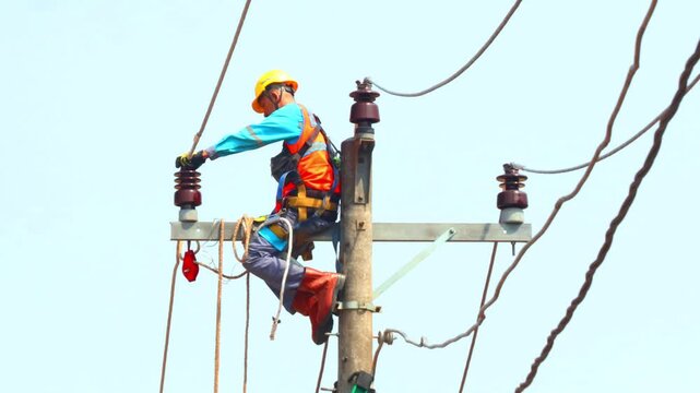 Electrician works on a tall power pole. Wearing safety gear and using tools, carefully handles cables and insulators on a three-phase power line. Focus demonstrates dedication and skill.