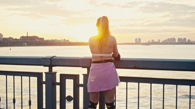 Tracking shot follows skater approaching city waterfront railing. Beautiful young woman stops to admire scenic river sunset relaxing after active street rollerblading session outside.